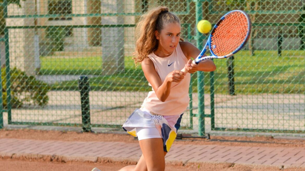 Energetic young female tennis player swings racket on outdoor clay court.